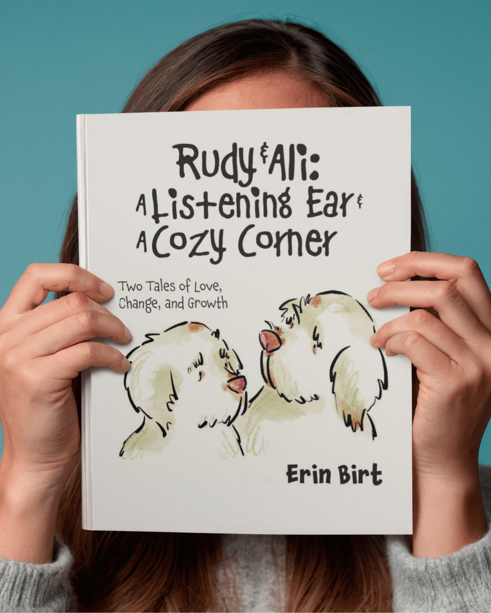 Hands of a woman holding the children’s book Rudy & Ali: A Listening Ear & A Cozy Corner by Erin Birt. The cover features two illustrated Clumber spaniels facing each other against a simple teal background.