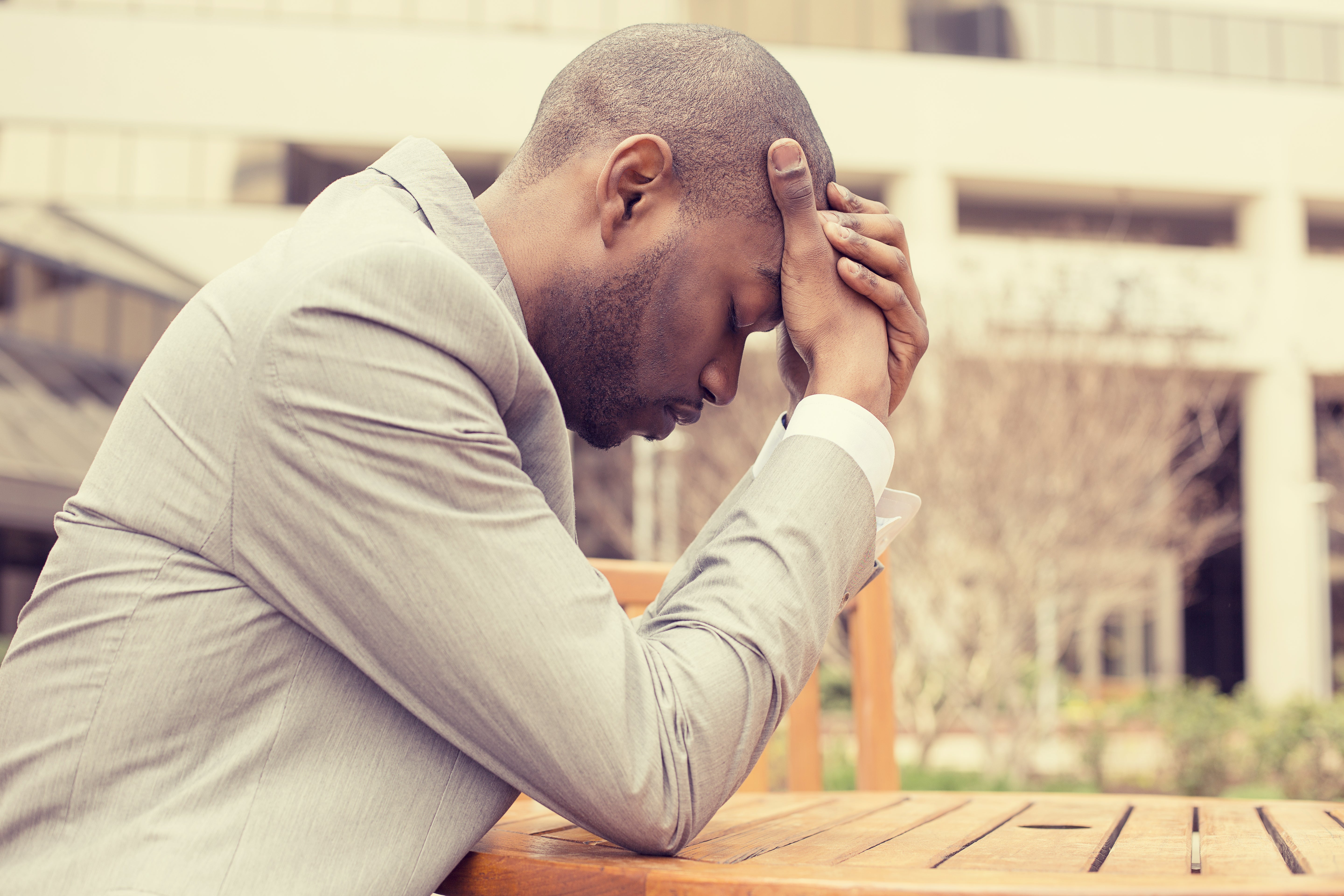 Distressed man sitting at a table outdoors with his head resting in his hands.