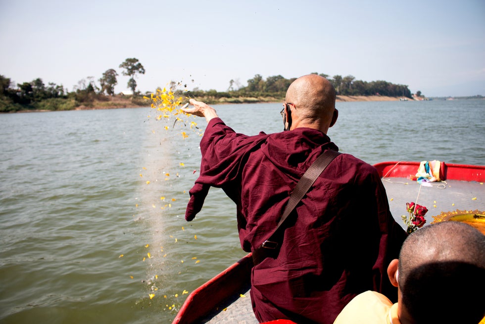 Man in monk robe scattering ashes and flower petals into water at funeral ceremony.