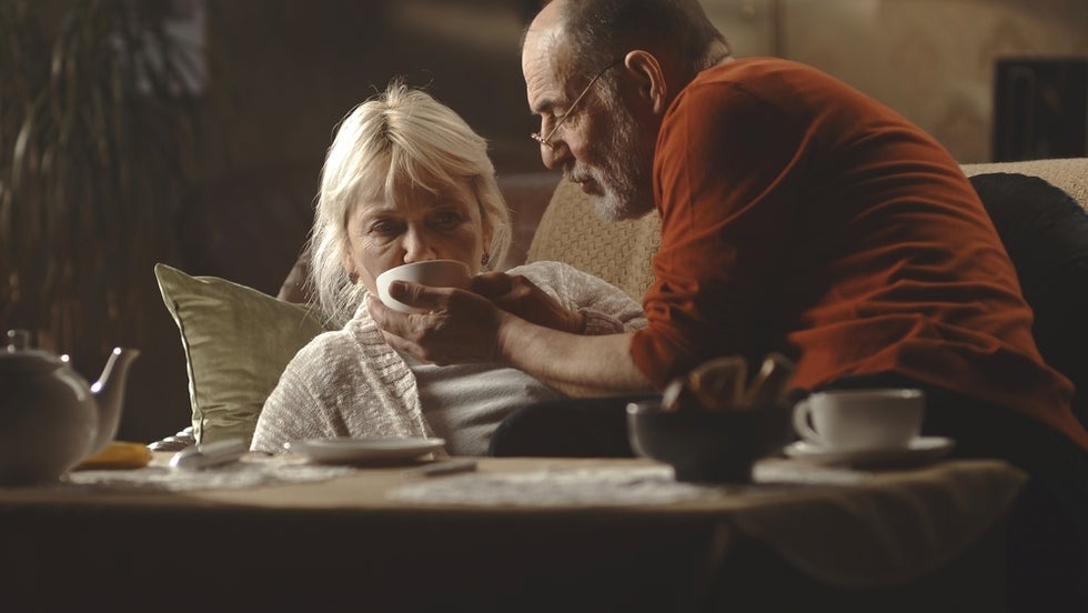 Elderly husband giving cup of tea to sick wife in living room at home