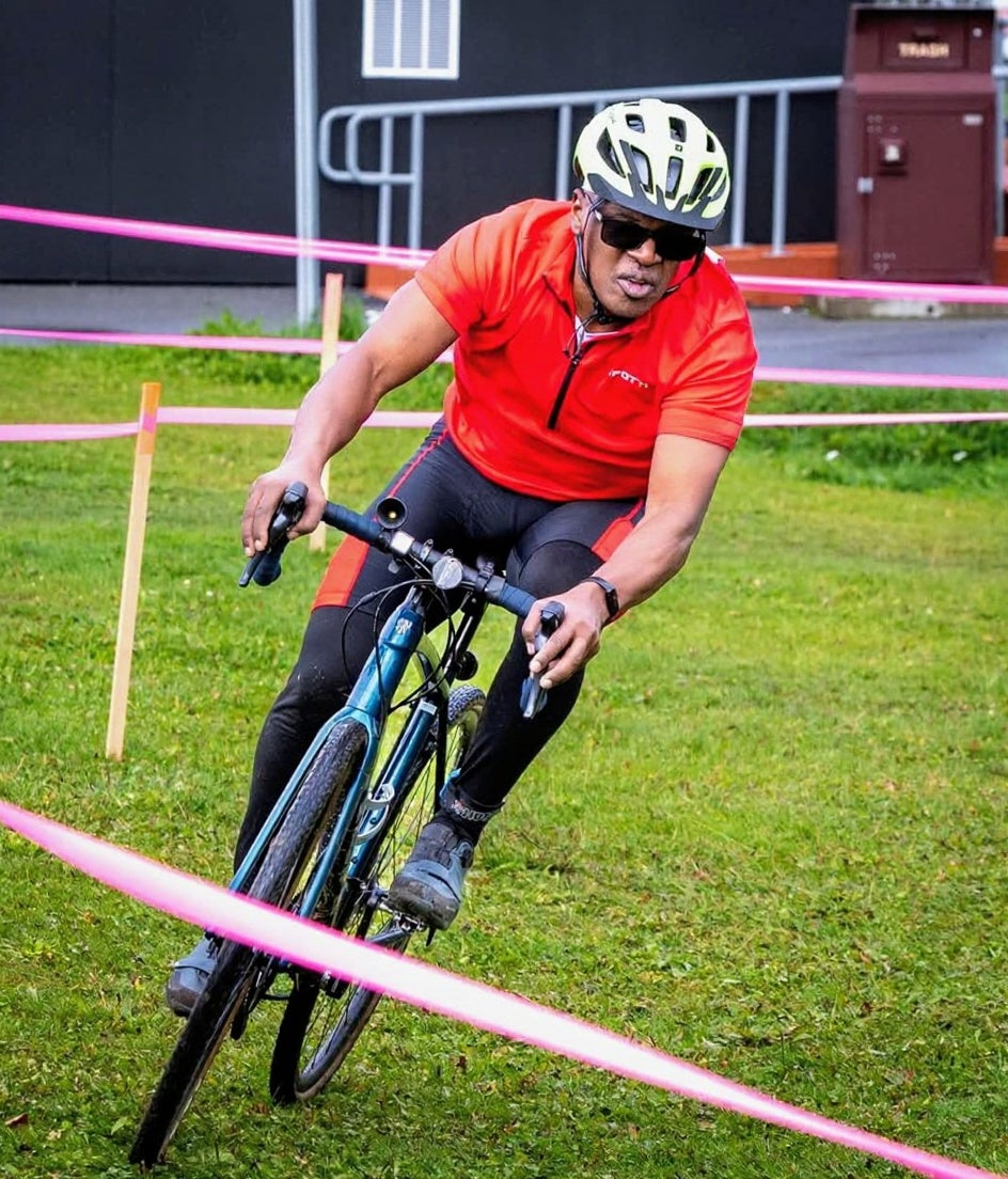 Cyclocross rider leaning hard into a grassy turn during Arctic Cross race, wearing a red jersey and helmet.
