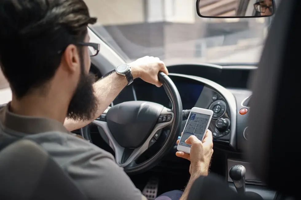  A man with a beard and glasses drives a car, holding a cell phone, highlighting Lyft ride share accident lawyers in New Jersey.
