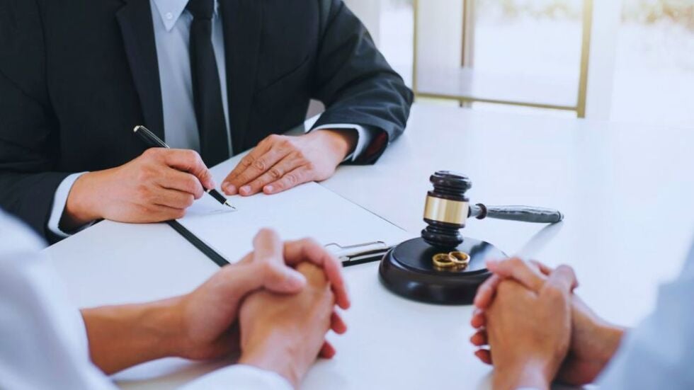 A divorcing couple sits across from a lawyer at a desk while the lawyer writes on legal documents, with a judge’s gavel and two wedding rings placed in front of them, symbolizing protecting a business during divorce discussions.