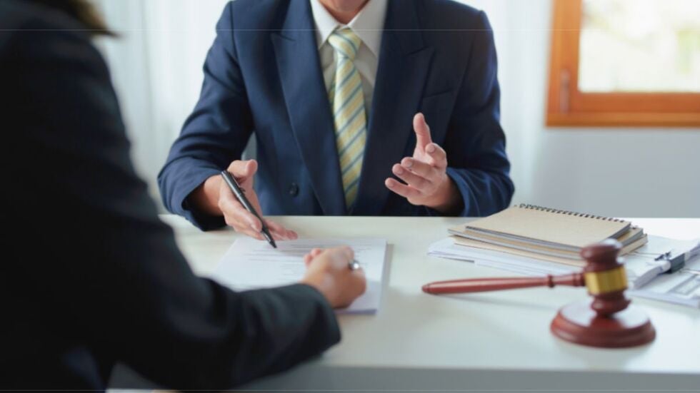 A man in a suit and tie sits at a desk with a woman, discussing legal matters related to enforcement.