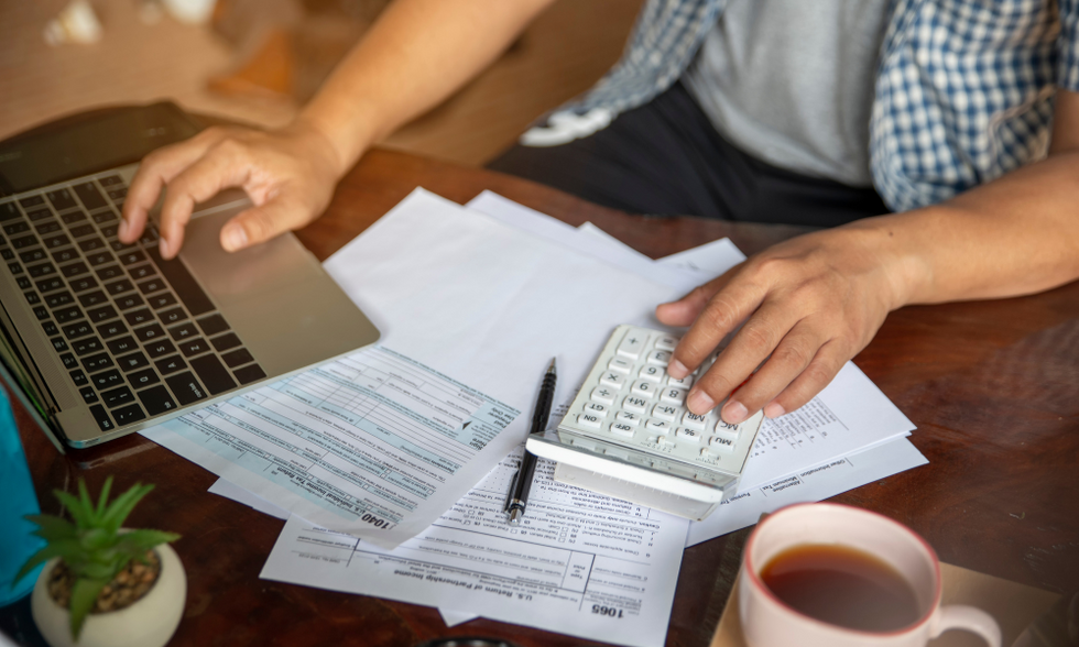 A man sitting in front of a laptop, a stack of bills, a calculator, and a cup of coffee, working on his budget.