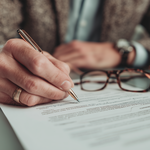 Close Up Picture of two hands, one hand is holding a pen and signing a document. Glasses are in front of the pen. 