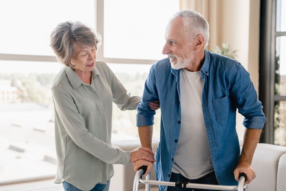 An elderly woman is helping an elderly man with a walker.
