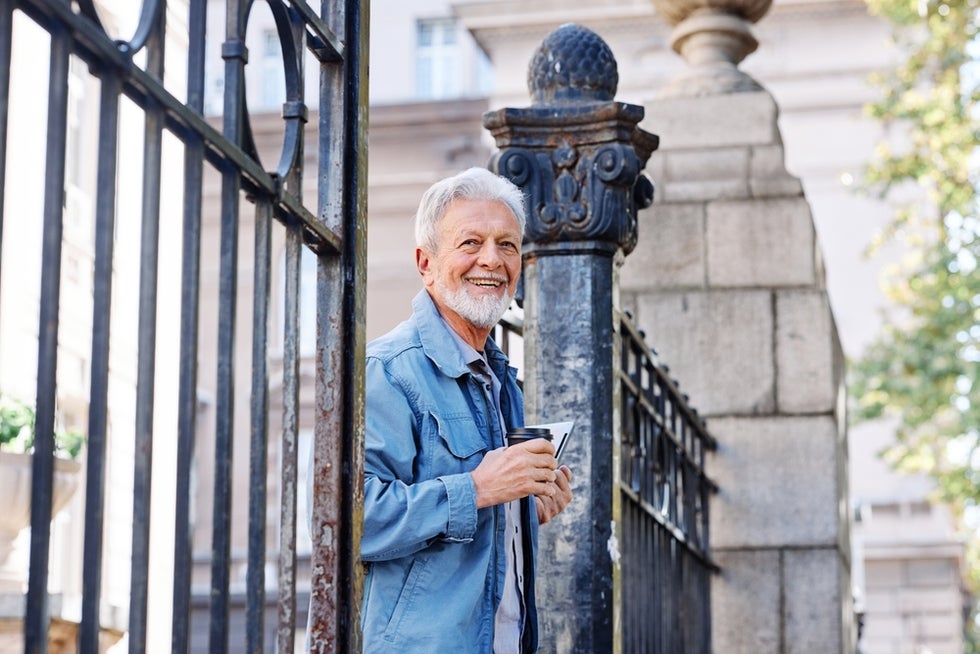 Smiling senior man stands at gate to college campus with coffee and laptop in hand.