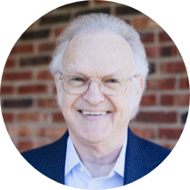 An elderly man with white hair and glasses, smiling, wearing a navy blazer and white shirt, standing in front of a brick wall.