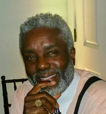 Homme noir souriant aux cheveux grisonnants et à la barbe, portant une chemise rose, des bretelles noires et une bague.